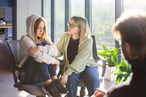 “A young child sitting alone, looking down and withdrawn, while his mother leans toward him with a concerned and caring expression, showing support and attention.”